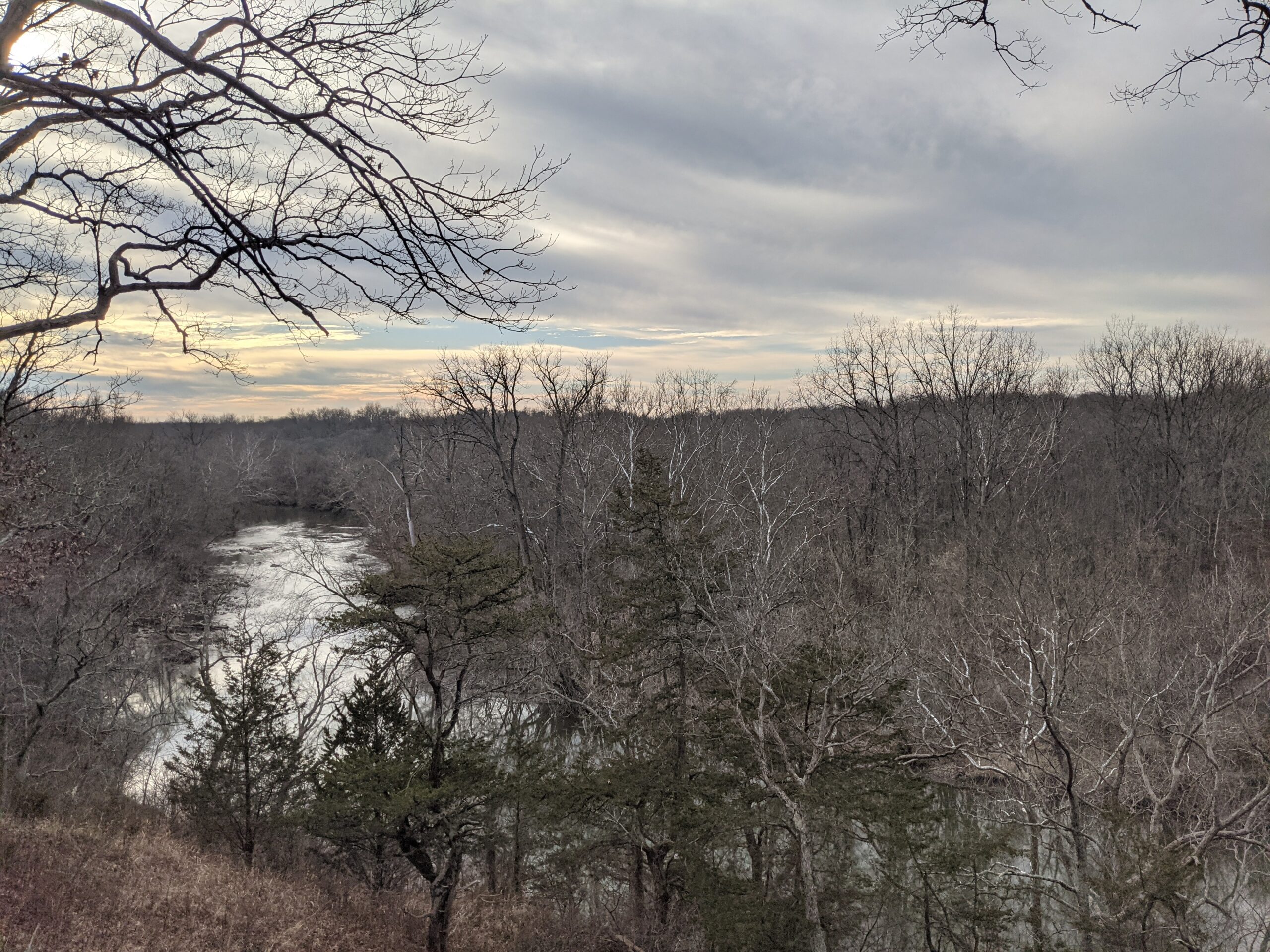 Zoomed out view of a landscape. There is a semi frozen creek running through a hilled landscape. There are barren trees on either side of the creek with a cloudy sky in the background.