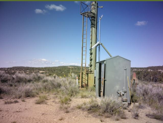 An orphan well comprised of a tall yellow pipe extending from beneath the ground to tens of feet in the air. Standing beside the pipe is a weathered grey control box. 